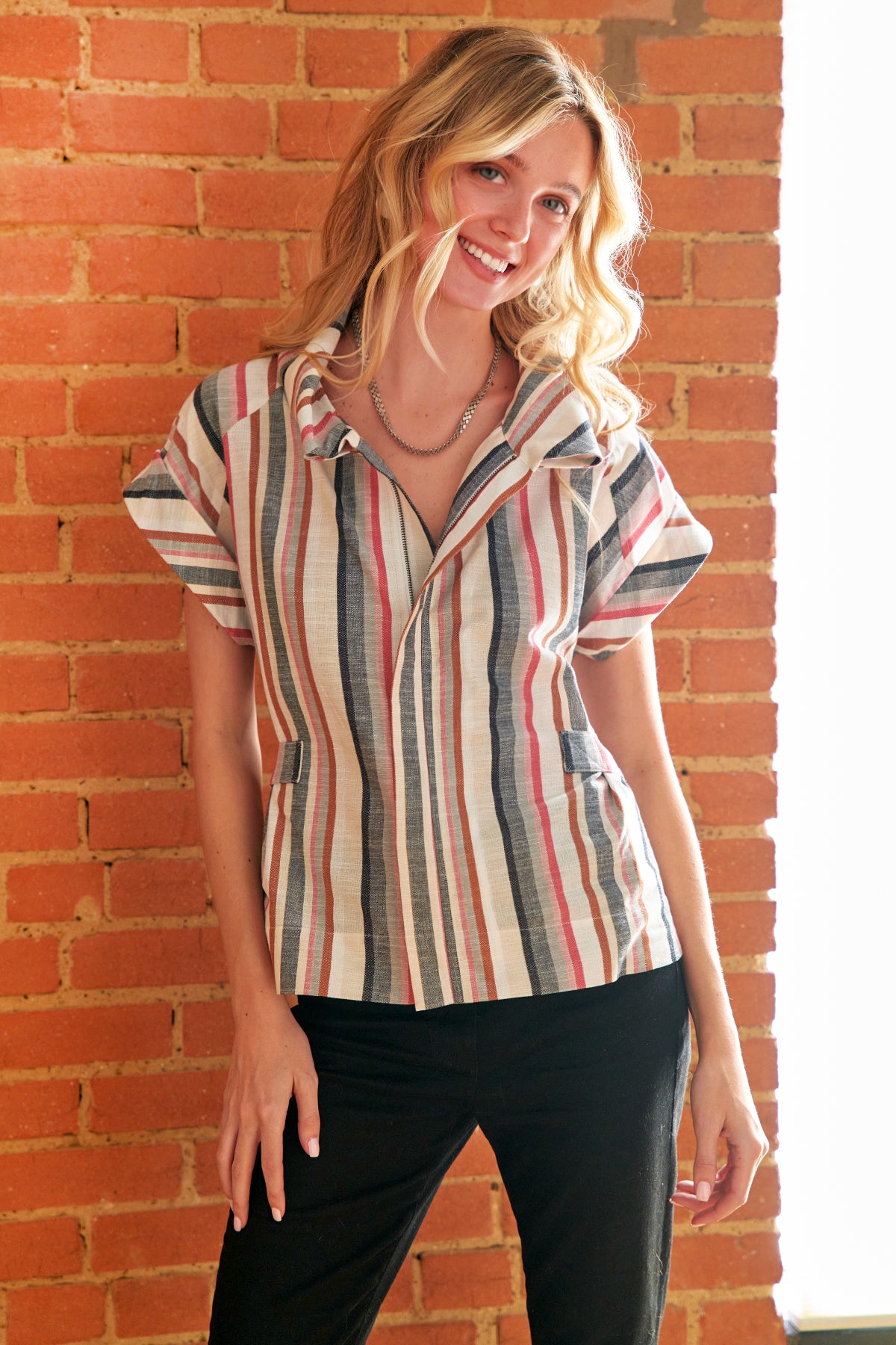 A woman wearing a short-sleeve striped button-up shirt with vertical stripes in beige, red, blue, and gray, paired with black pants, standing against a brick wall.