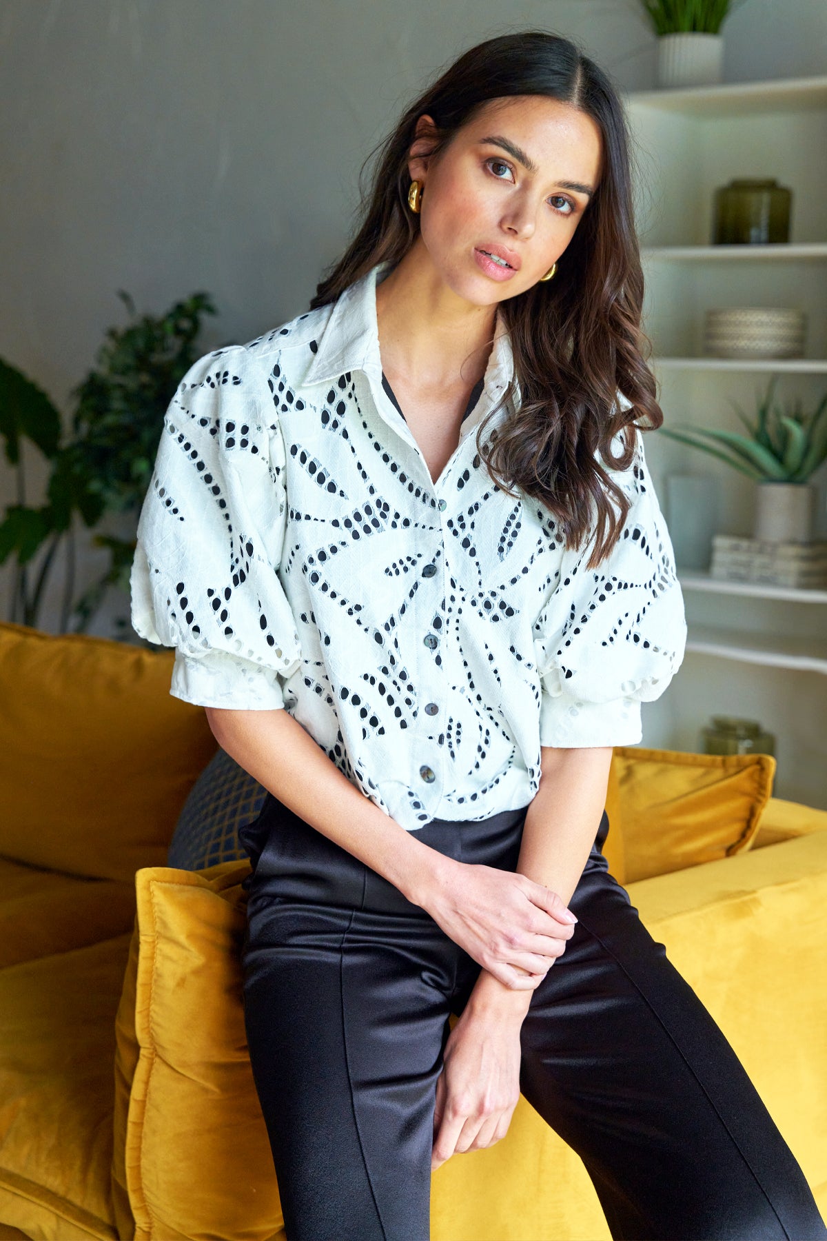 Woman sitting on a yellow couch wearing a light blue blouse with black patterns and dark pants.