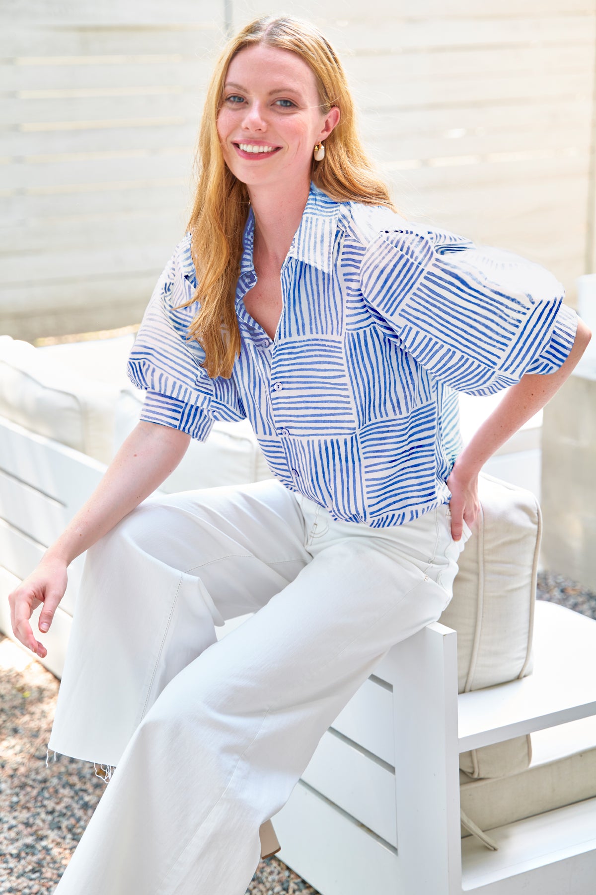 Woman wearing a blue and white striped shirt and white pants sitting on a white bench.