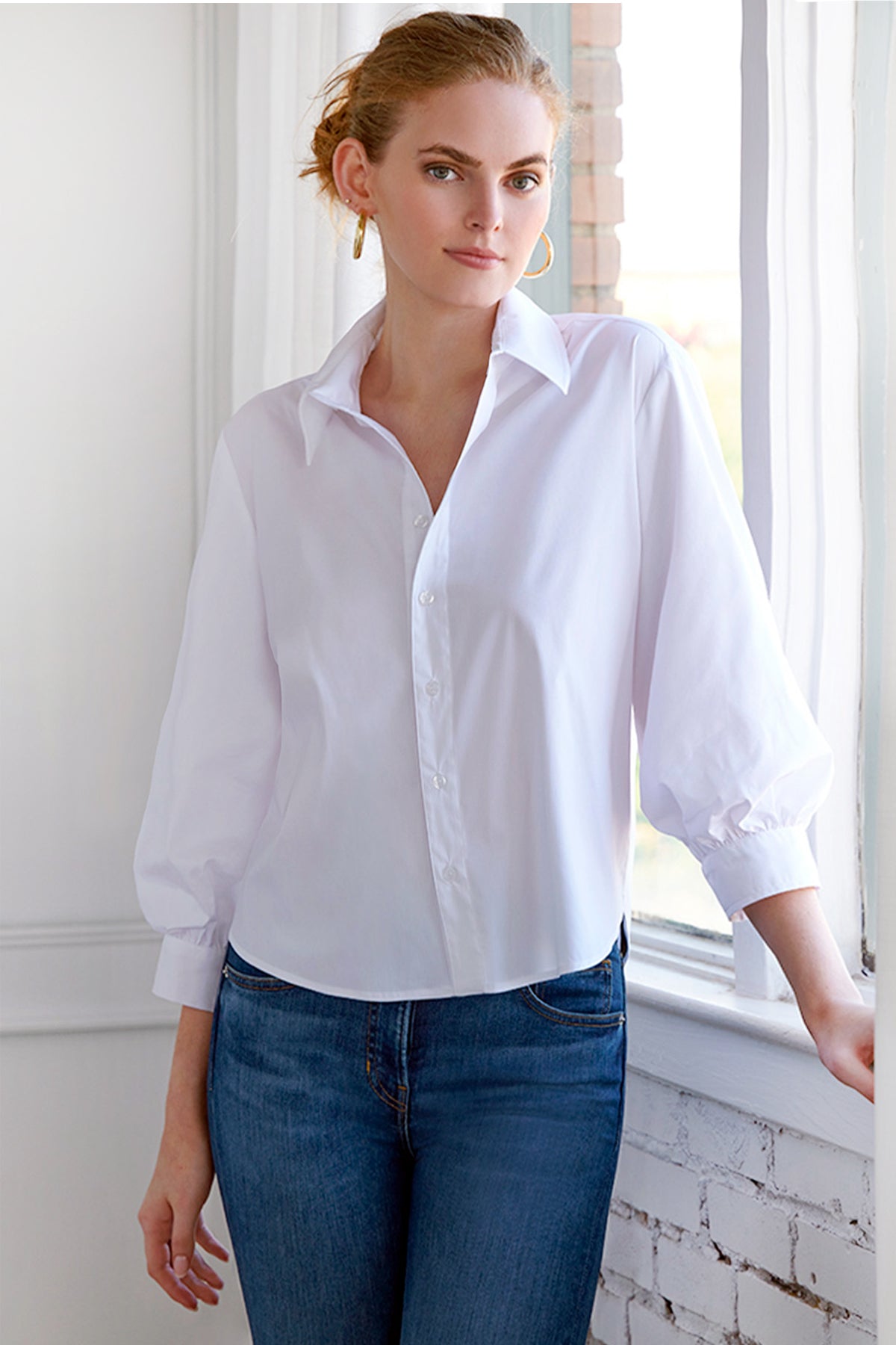 Woman wearing a white shirt and blue jeans standing by a window.