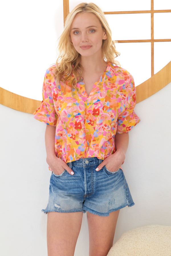 A model wearing the Finley Crosby blouse, a short puff sleevd v-neck blouse with an abstract pink and orange capri floral pattern.
