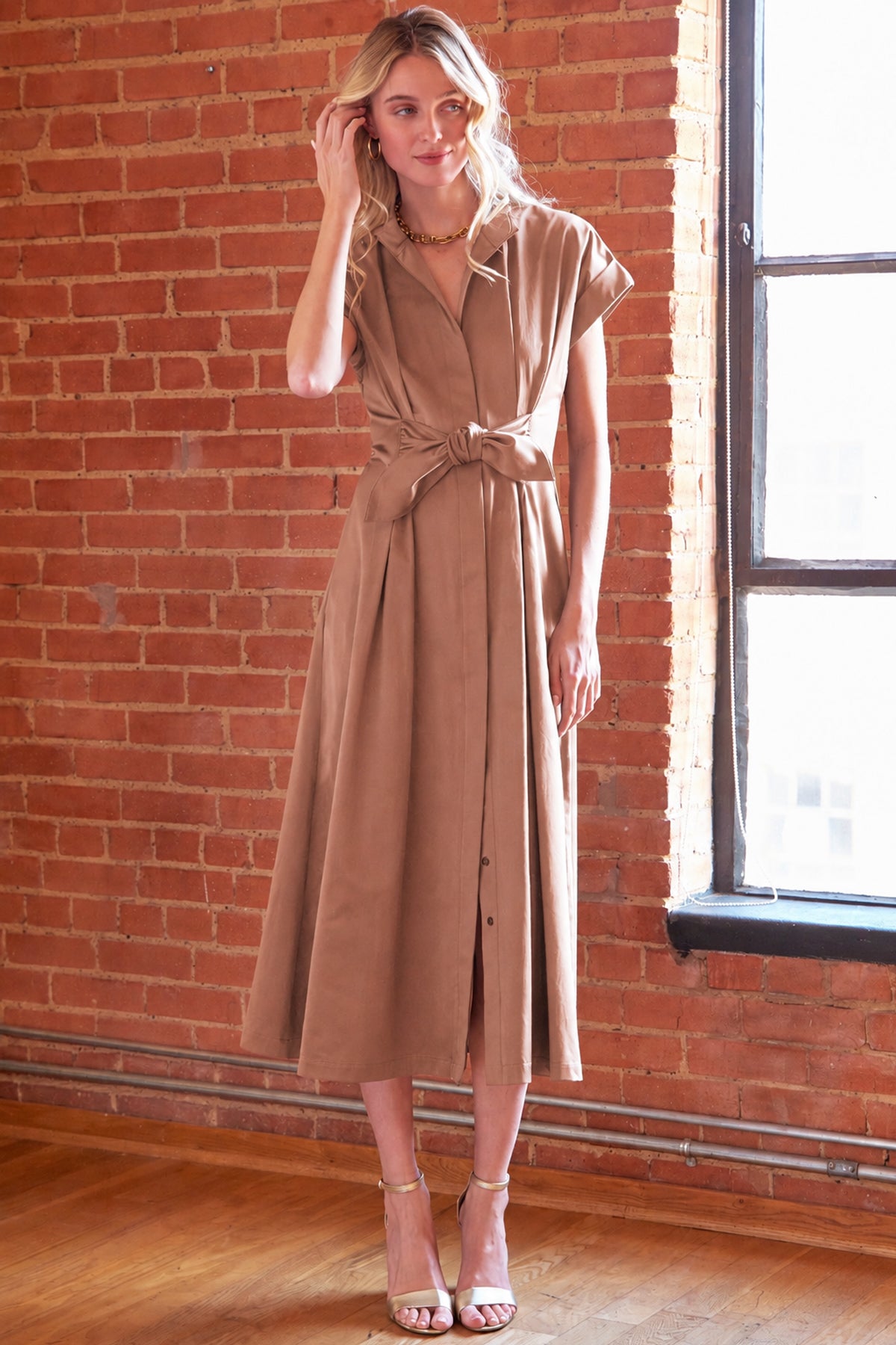 A woman models a taupe brown midi shirt dress with short sleeves, a button-down front, and a tied waist, standing in front of a brick wall next to a window.