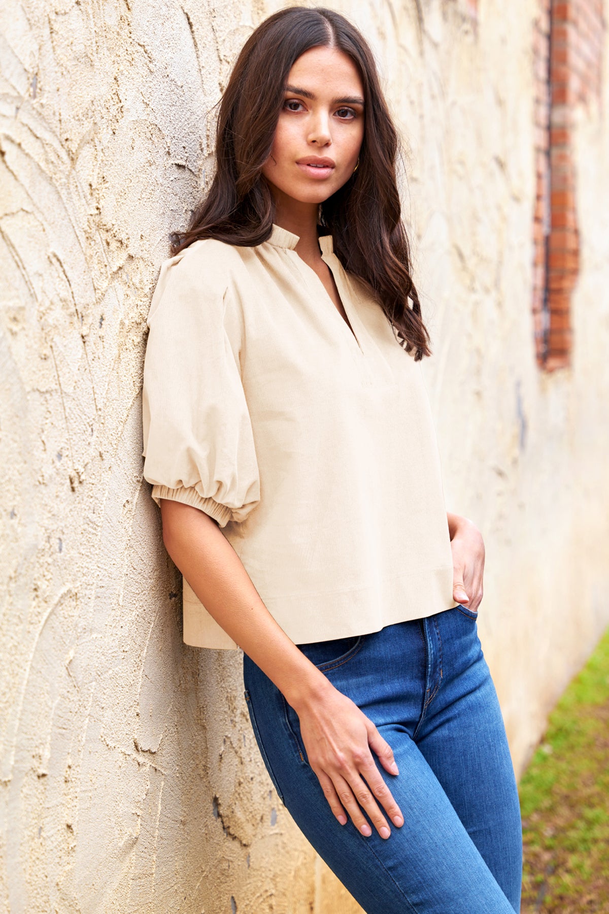 Woman wearing a beige blouse and blue jeans leaning against a stone wall.