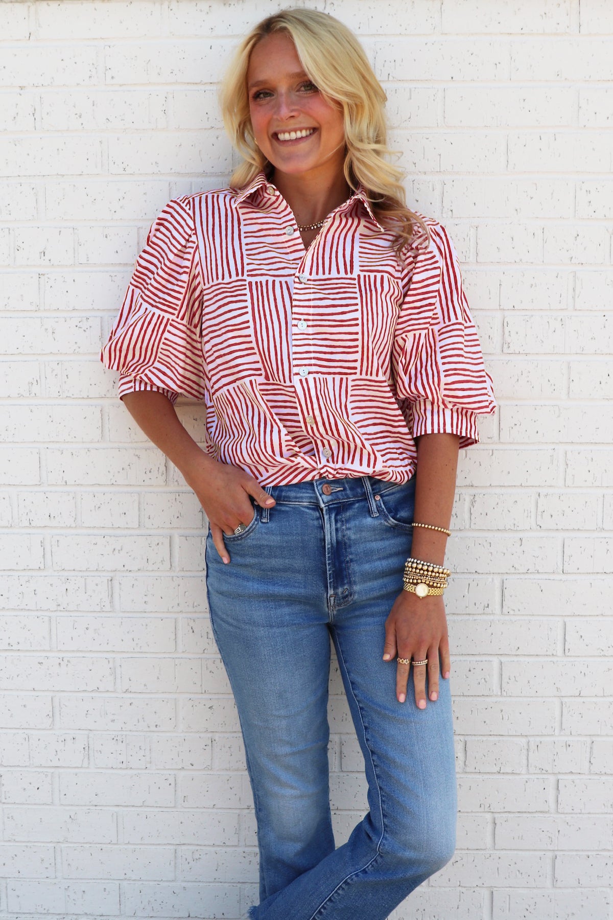 Woman wearing an orange and white patterned blouse and blue jeans against a white brick wall.
