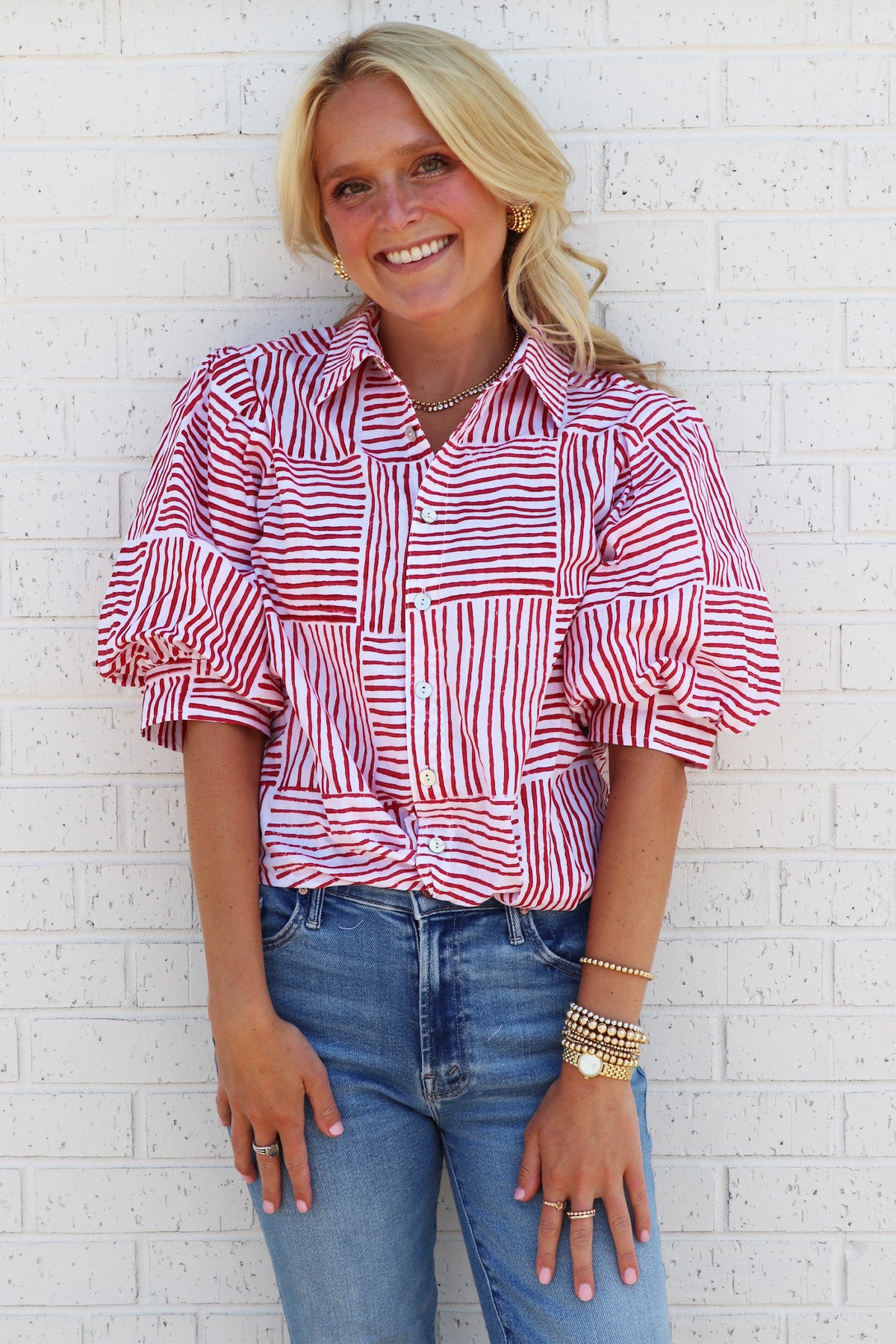 Woman wearing a red striped puff sleeve womens blouse and blue jeans against a white brick wall.