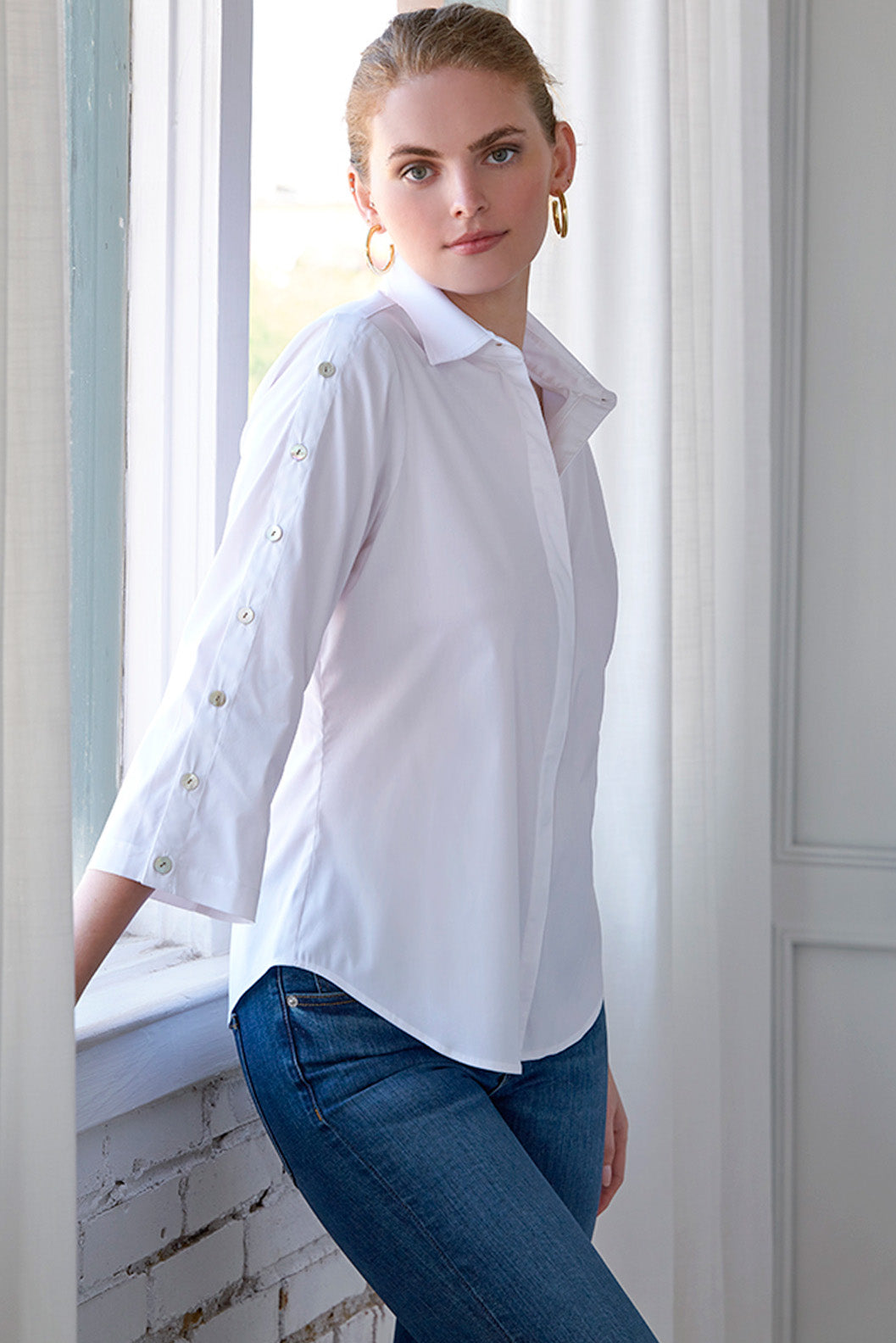 Woman wearing a white blouse and blue jeans standing by a window.