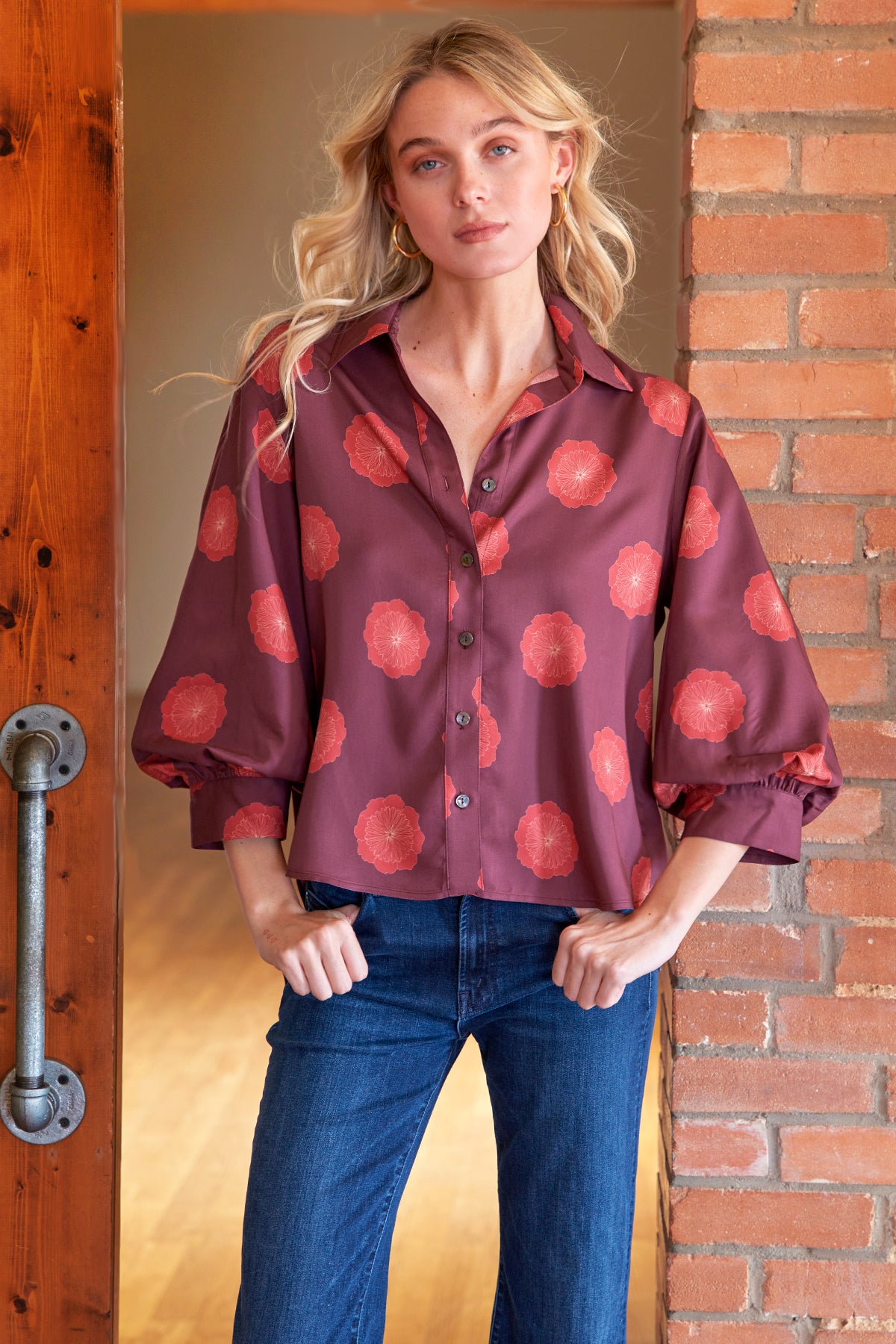 A woman wearing a mauve button-up shirt with a coral-red floral pattern and blue jeans, standing in a doorway between a wooden door and a brick wall.