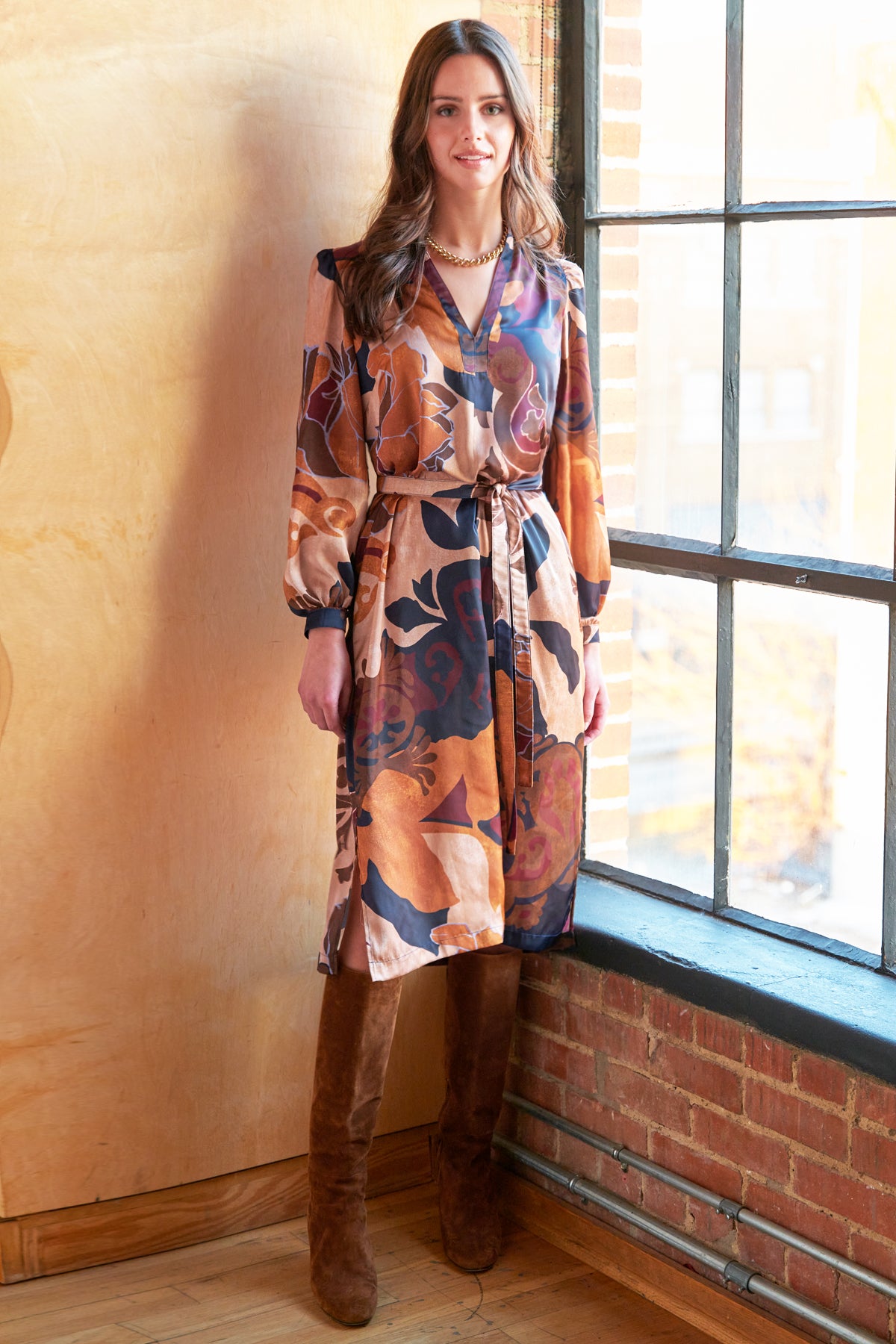 Woman wearing a patterned dress standing by a window with a brick wall background