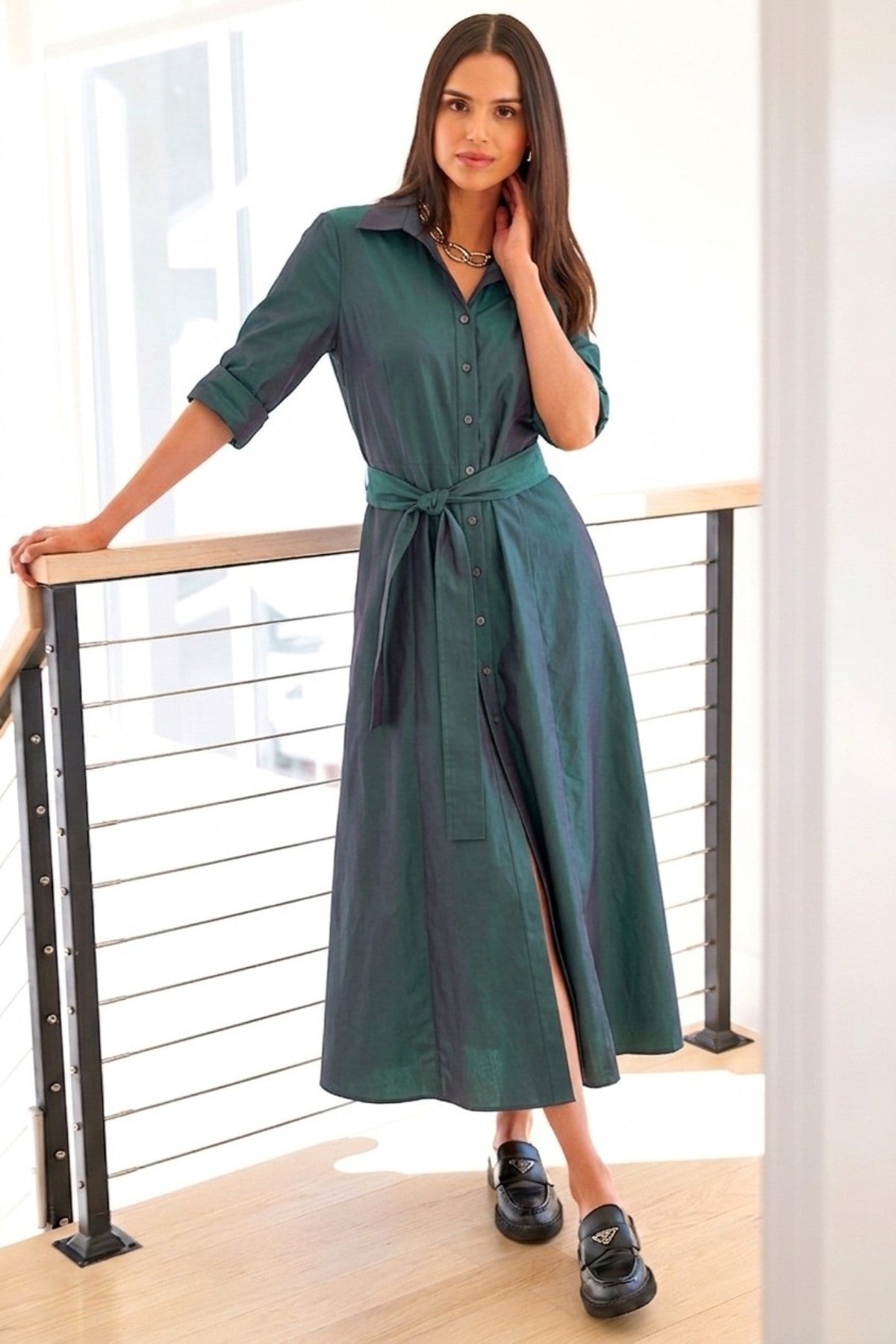 A woman models a dark green midi shirt dress with a tie waist belt, standing on a wooden deck next to a modern railing.