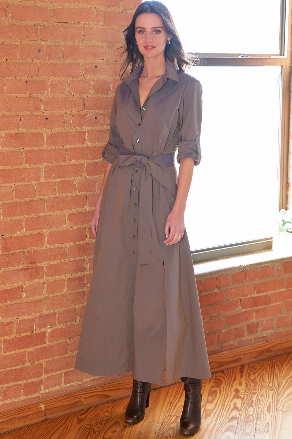A woman models a taupe-brown midi shirt dress with rolled sleeves, a button-down front, and a matching waist tie, styled with black ankle boots in front of a brick wall and window.