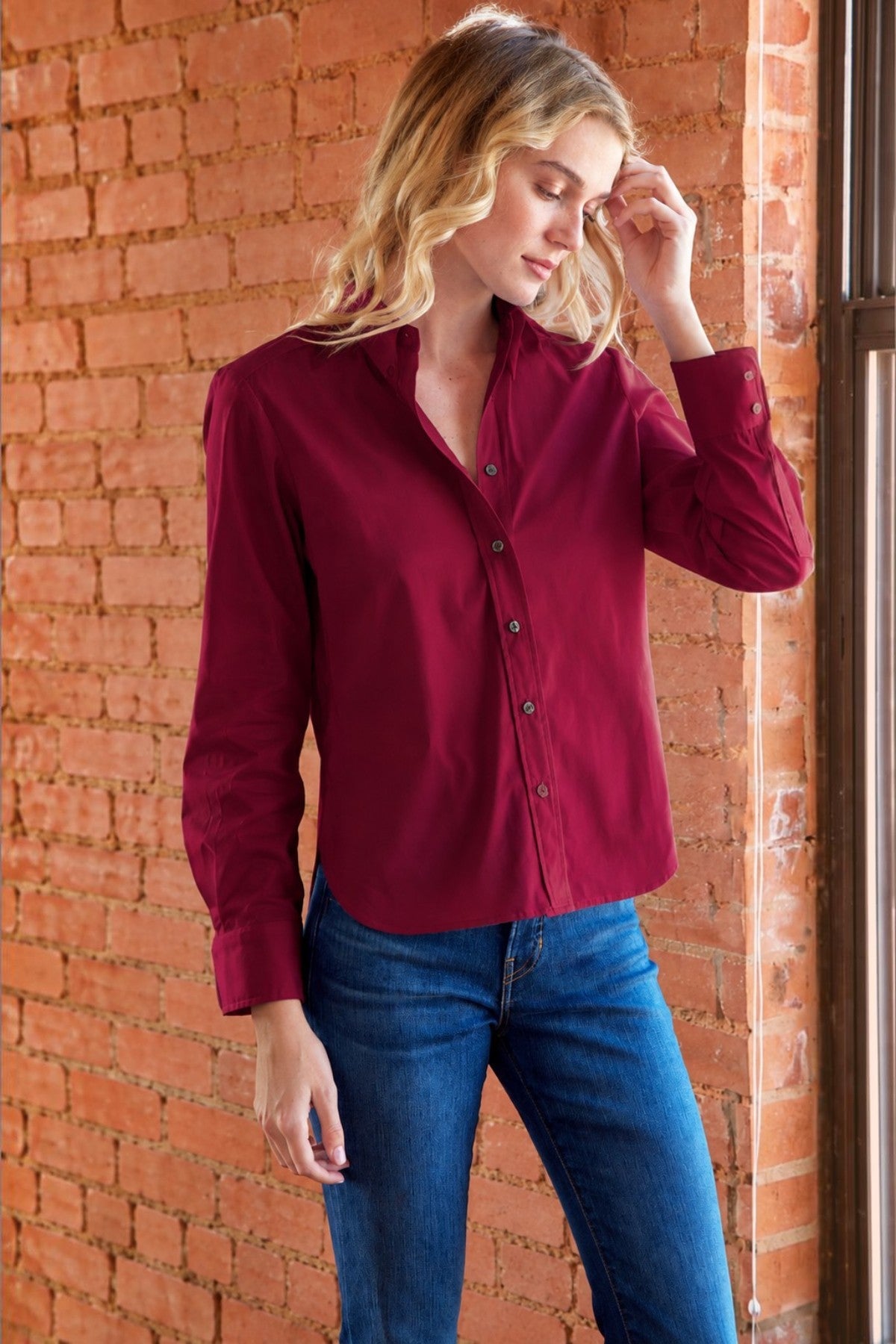 A woman wearing a burgundy button-down shirt and blue jeans stands against a red brick wall.