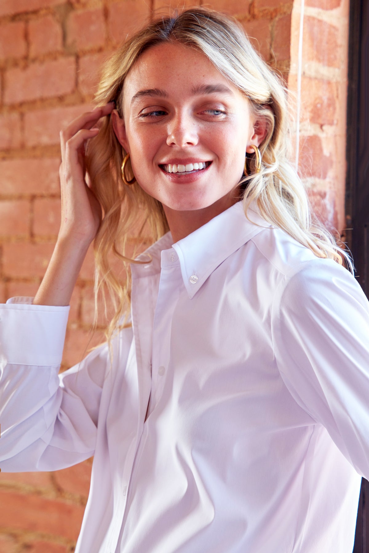 A smiling blonde woman wearing a white button-down shirt and gold hoop earrings, standing in front of a warm brick wall.