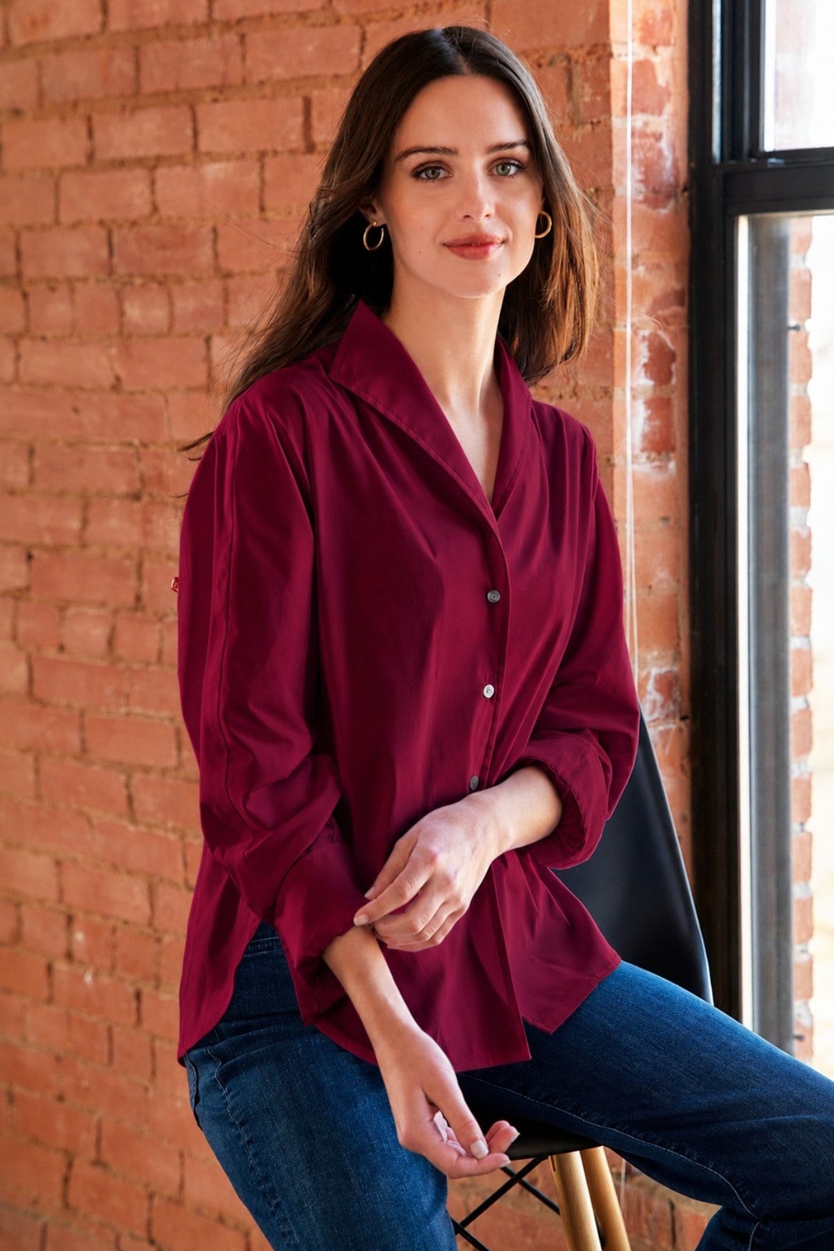 A woman wearing a long-sleeve burgundy button-up shirt and blue jeans, sitting on a black chair in front of a brick wall next to a window.