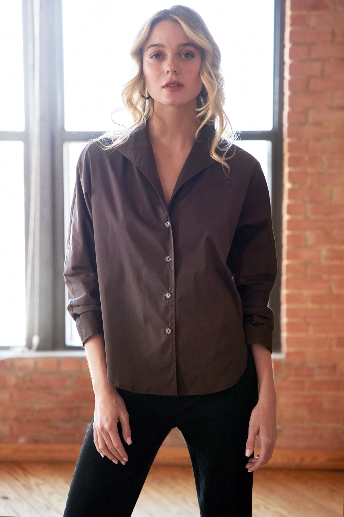 Woman wearing a brown button-up shirt in a room with brick walls and large windows.