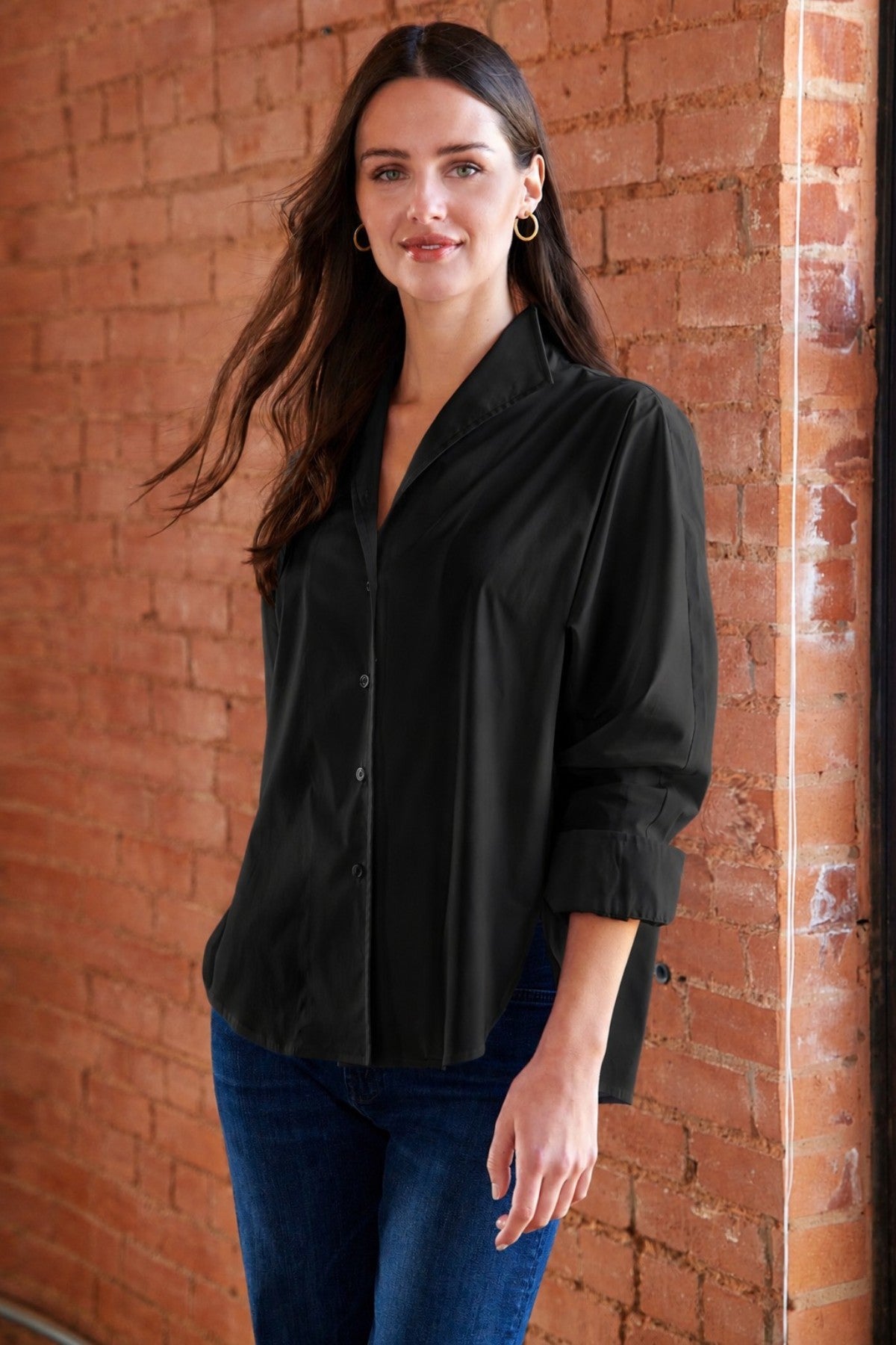 A woman wearing a black button-up shirt and blue jeans stands against a red brick wall.