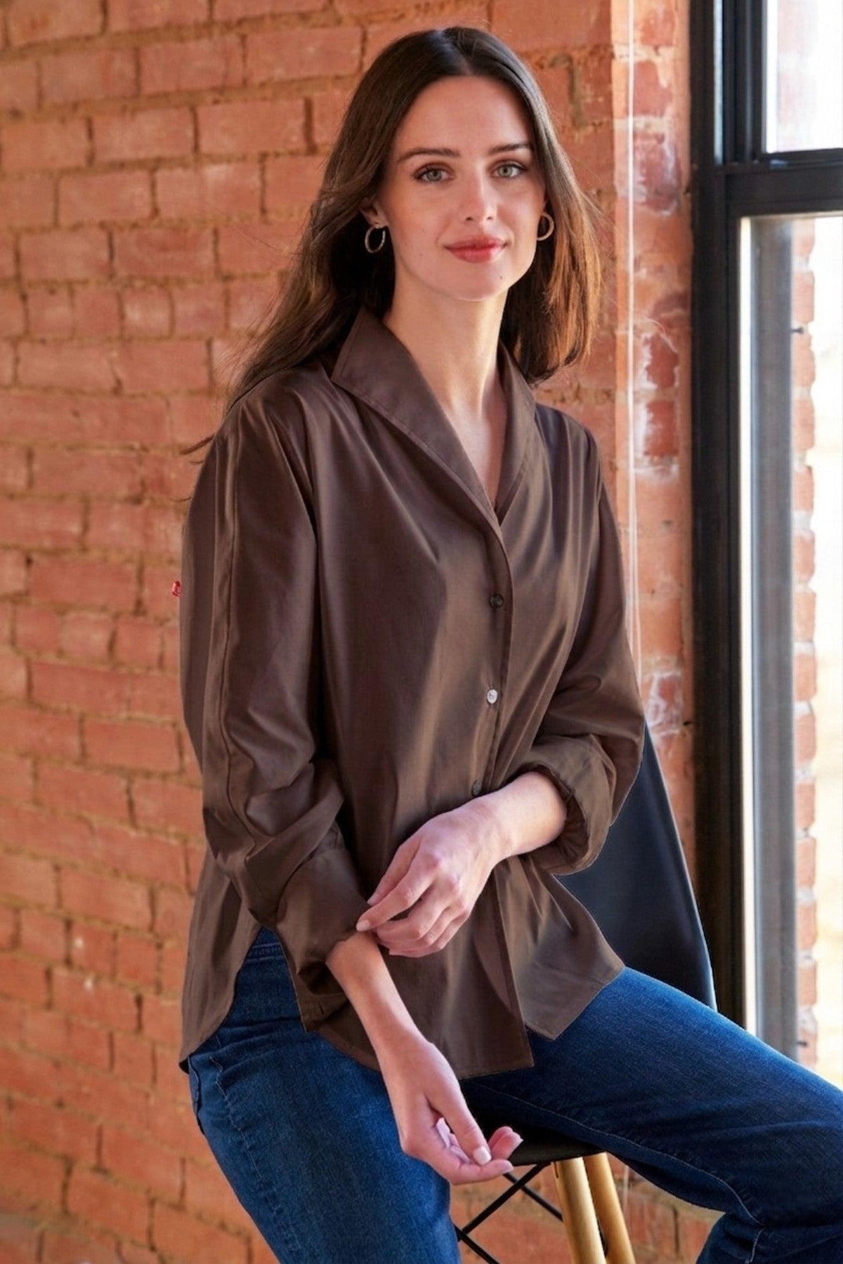 A woman wearing a brown satin-like button-up blouse and blue jeans, seated on a stool in front of a brick wall and window.
