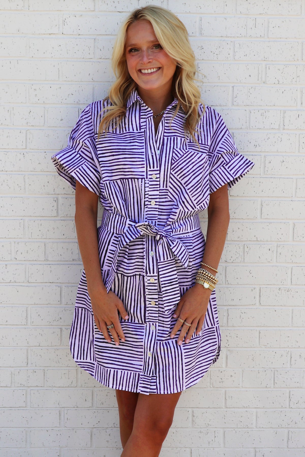 Woman wearing a purple and white striped dress against a white brick wall.