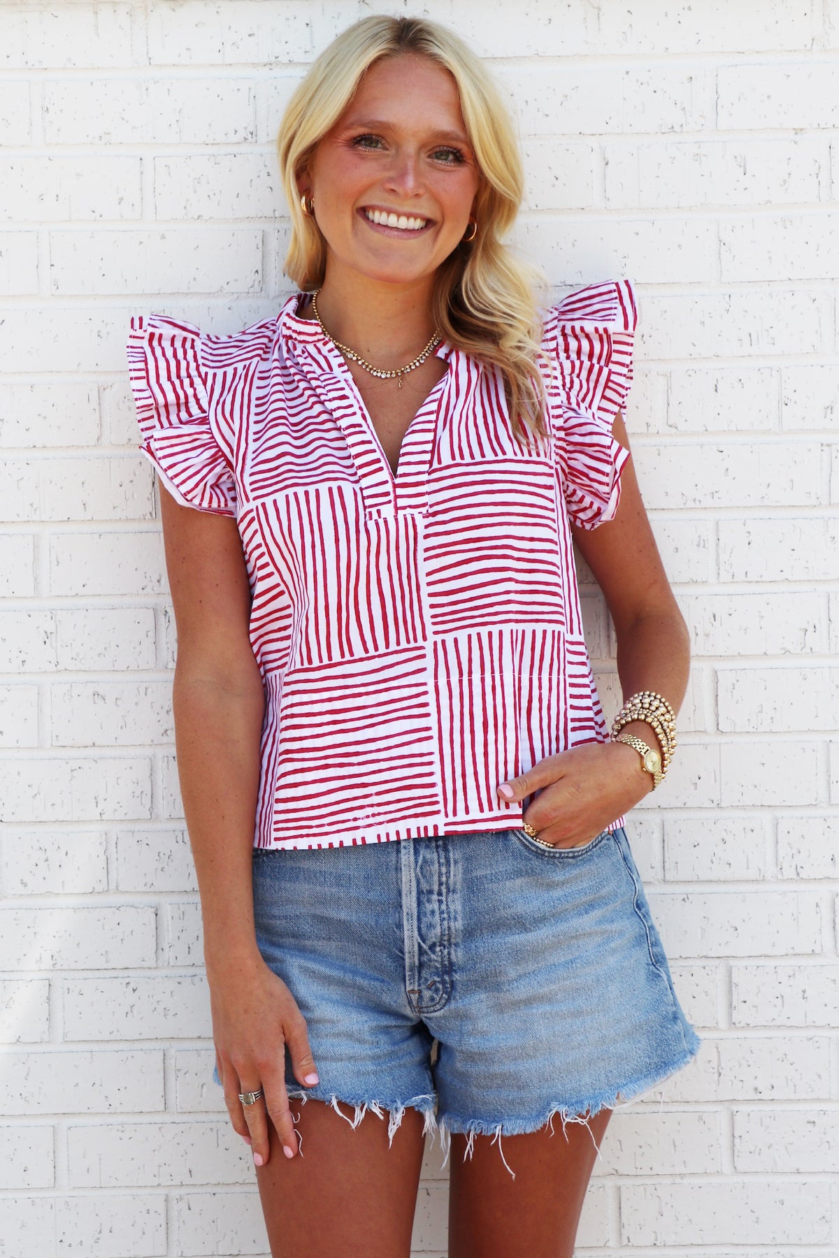 Woman wearing a red and white striped shirt and denim shorts against a white brick wall.