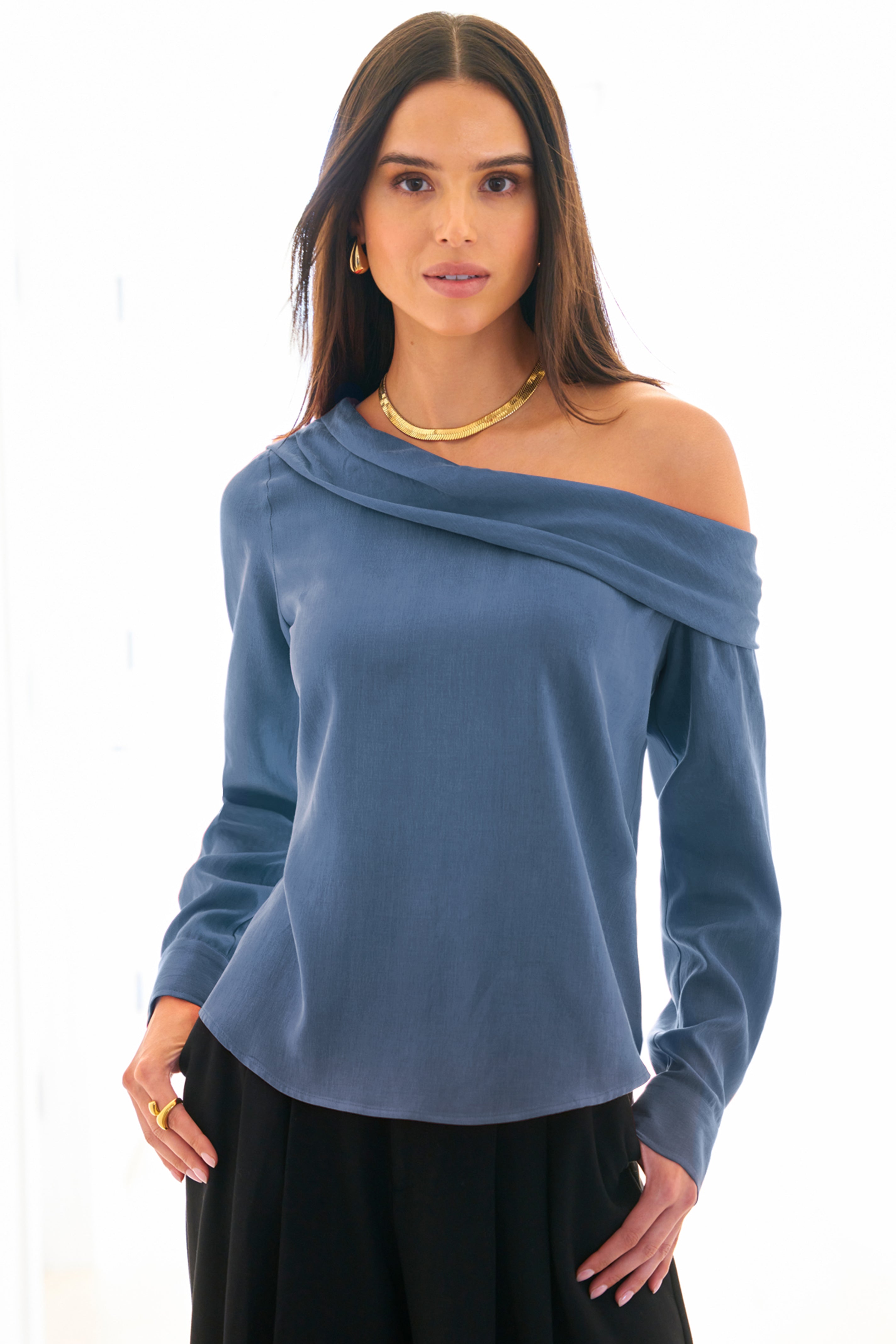 A model wearing the Dani shirt, featuring long sleeves, an off-shoulder foldover neckline, cupro cotton fabric in dark teal, and a relaxed shape.
