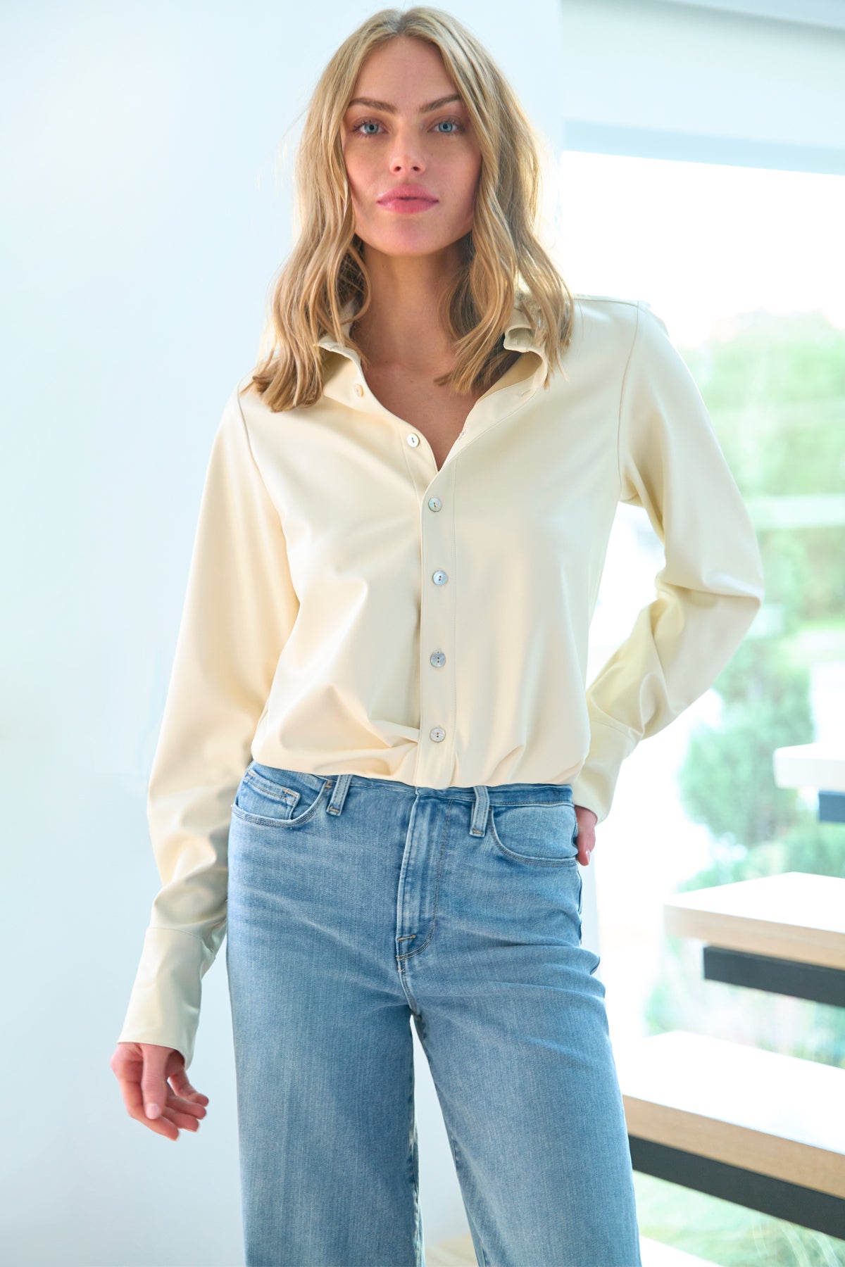 A model wearing the Moxie shirt, made from vegan leather in ivory, featuring a front-twist elasticized hem, cropped fit, and long sleeves.
