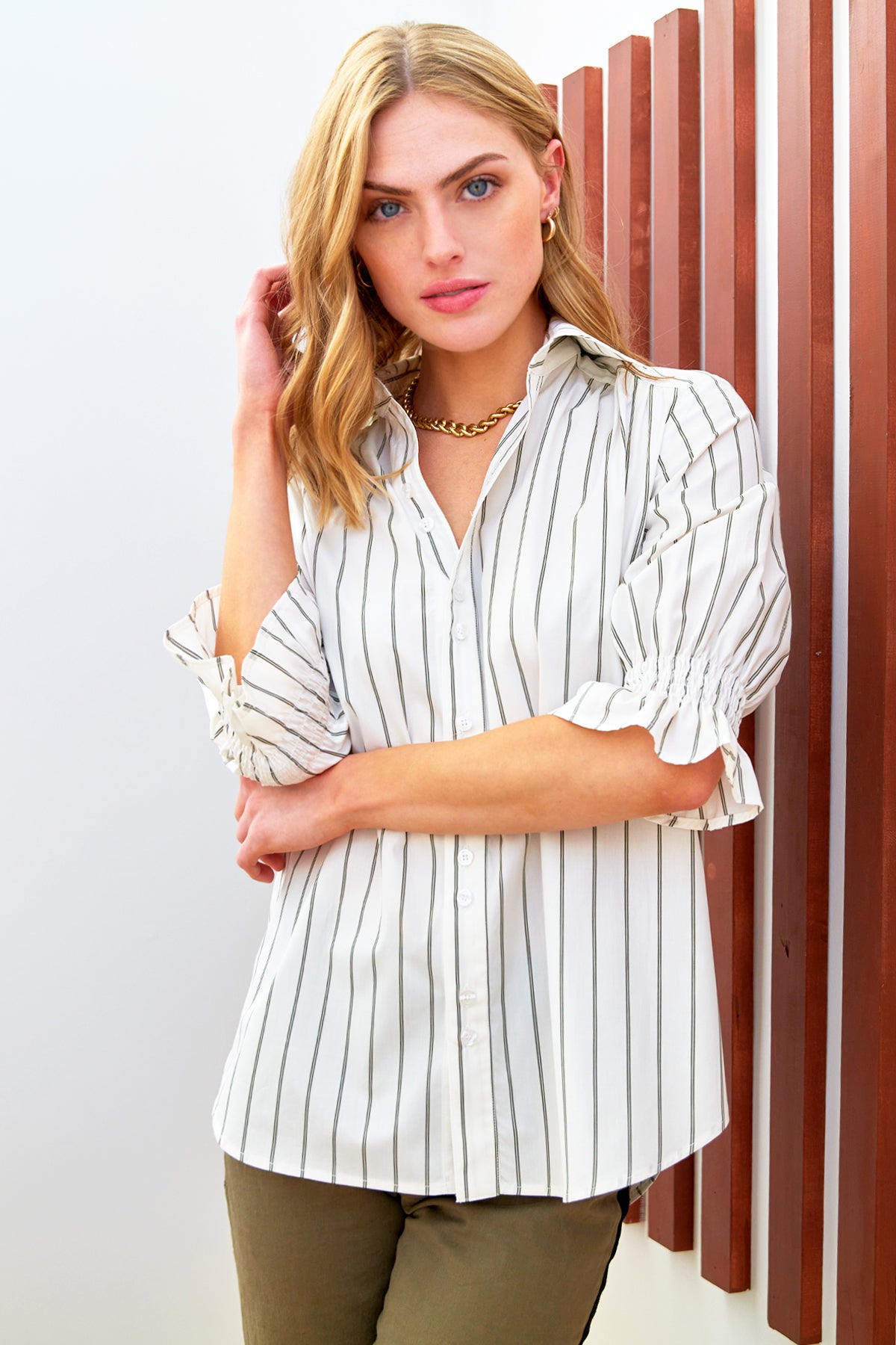 A model wearing the Finley Sirena blouse, a button-down cotton blouse with short puff sleeves and an olive pinstripe pattern.