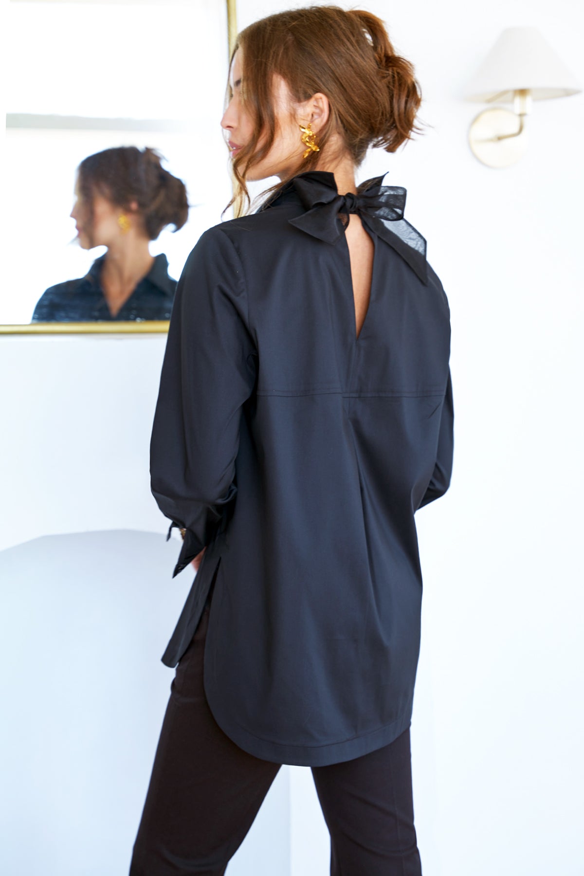 A model showcasing the Sylvie shirt with a cutout back self-tie, black organza bow, relaxed contour, and long sleeves.
