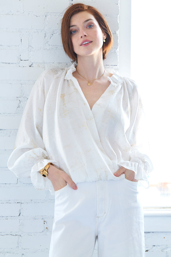 A model wearing the Finley Brette blouson, a ruffle trim white blouse with a v-neckline and a metallic gold texture.
