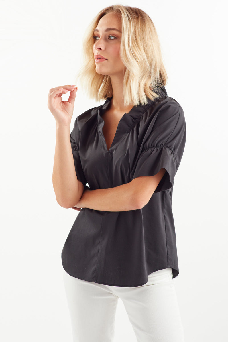 A blonde fashion model wearing the Finley Crosby shirt, a black poplin blouse with a ruffle collar and French sleeve detail.